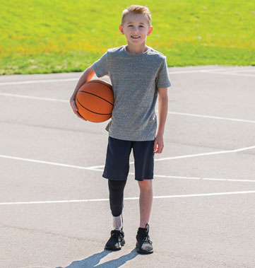 Un garçon amputé d’une jambe se tient debout sur un terrain de sports avec un ballon de basketball sous son bras.