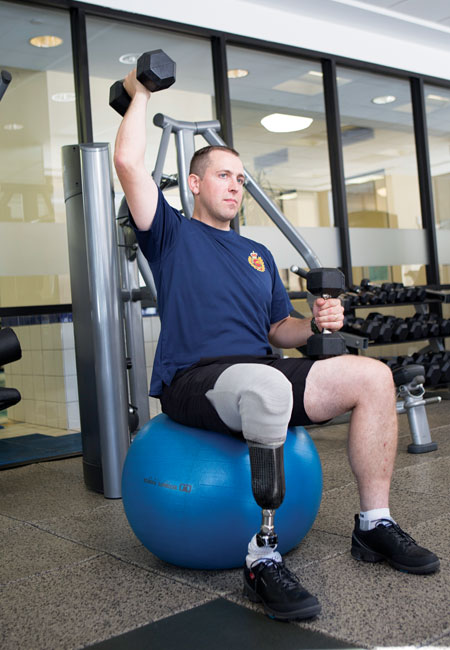 Un vétéran de la guerre d’Afghanistan amputé d’une jambe est assis sur un ballon d’exercice et soulève des poids avec ses bras dans une salle de sport.