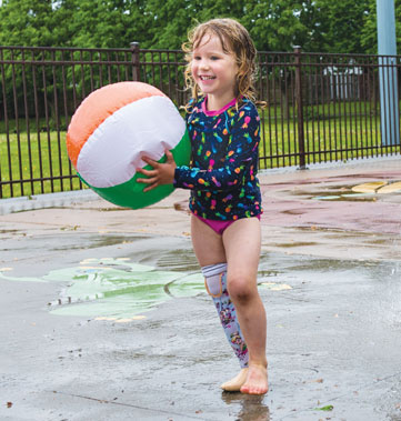 Une fillette amputée à la jambe droite tient un ballon de plage en jouant dans une aire de jeux d’eau.