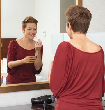 An adult female arm amputee looks in the mirror while applying lip gloss.
