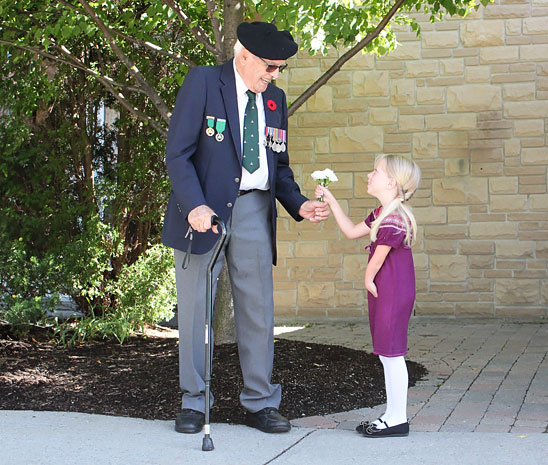 Une jeune fille qui a qui a une amputation au bras gauche tend un bouquet de fleurs à un ancien combattant de la Seconde Guerre mondiale qui est amputé d’une jambe. 