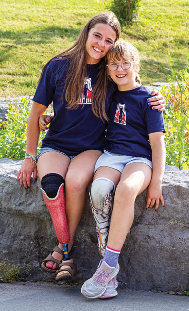 Une jeune femme et une petite fille, toutes les deux amputées d’une jambe, sont assises l’une à côté de l’autre sur une grosse roche, bras dessus bras dessous.