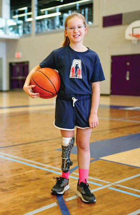 Une petite fille amputée d’une jambe tient un ballon de basketball en étant debout sur un terrain de basketball vide.