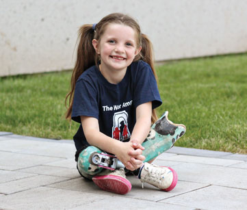 Une petite fille amputée des deux jambes est assise avec les jambes croisées sur le sol à l’extérieur.