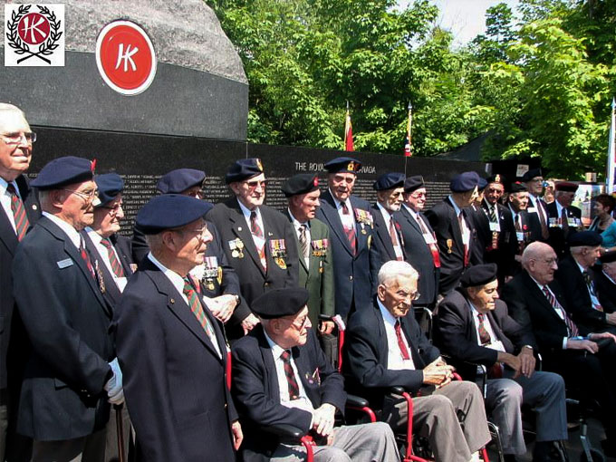 Plusieurs anciens combattants portant leur uniforme et leurs décorations militaires sont debout ou assis devant le mur commémoratif.
