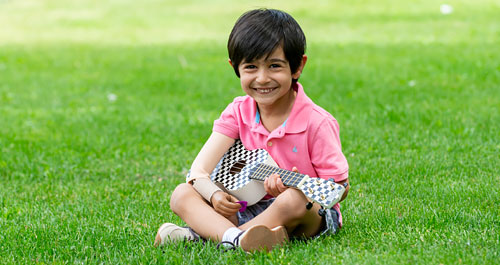 Un petit garçon amputé d’un bras est assis dans l’herbe et joue du ukulélé.